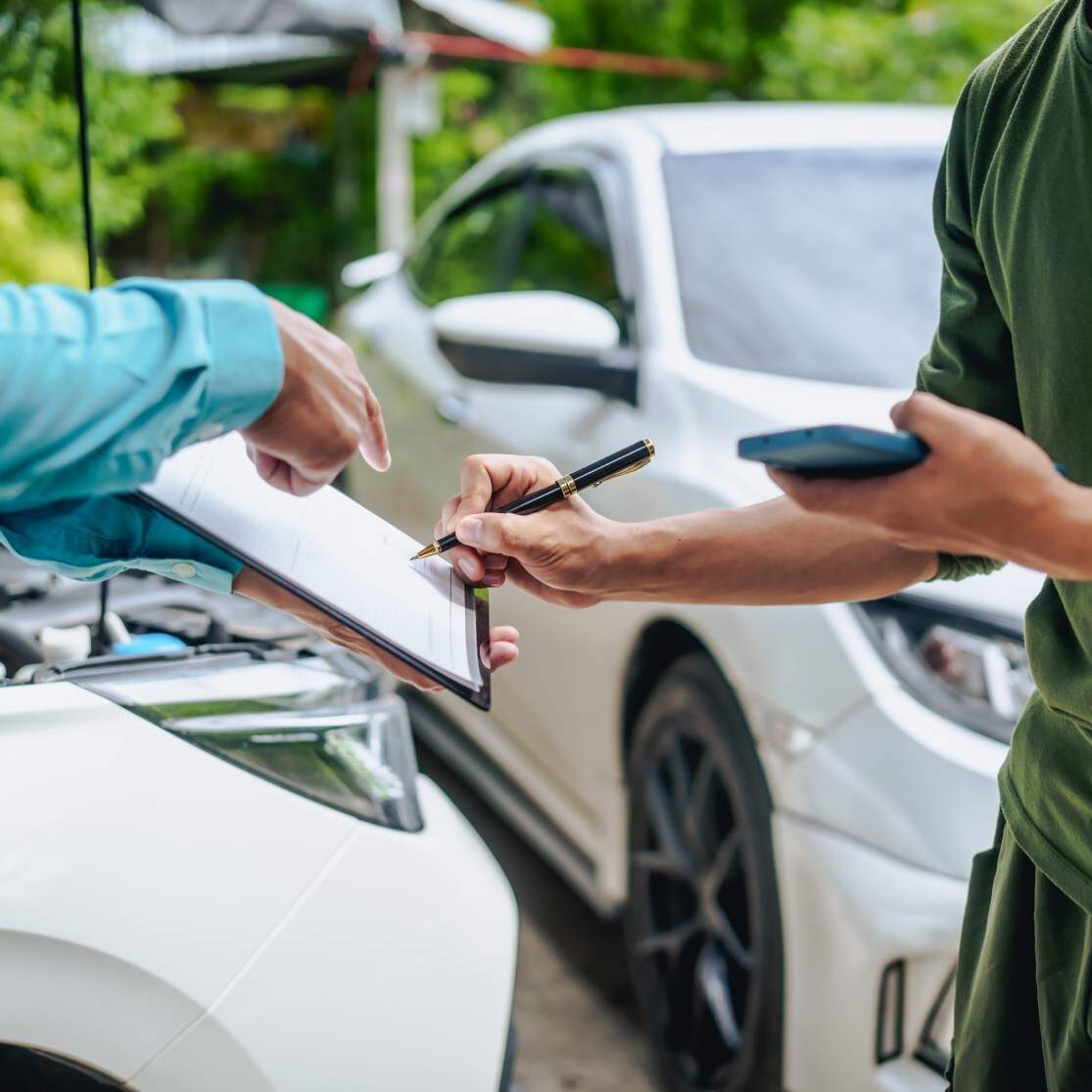 Asian clients engage with a car insurance agent beside a wrecked vehicle. They examine the policy details, assess the damage, and finalize paperwork related to the insurance claim and repairs.